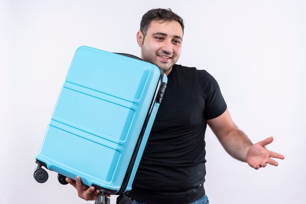 Homme jeune voyageur en t-shirt noir tenant valise à côté souriant joyeusement propagation avec bras sur le côté debout sur un mur blanc