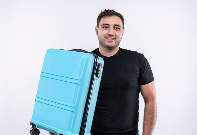 Homme jeune voyageur en t-shirt noir tenant valise heureux et positif avec sourire sur le visage debout sur un mur blanc