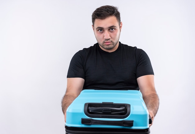 Homme jeune voyageur en t-shirt noir tenant une valise avec un visage sérieux debout sur un mur blanc