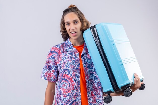 Homme jeune voyageur avec valise bleue à la recherche de sourire confiant avec visage heureux prêt à voyager debout