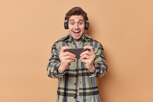 l'homme joue à un jeu vidéo mobile tient le smartphone horizontalement porte des écouteurs sans fil porte une chemise à carreaux isolée sur beige. Concept de personnes et de technologie