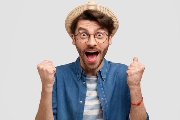 Homme joyeux agronome serre les poings, ouvre largement la bouche, s'exclame avec bonheur, porte un chapeau de paille et une chemise en jean décontractée, isolé sur un mur blanc