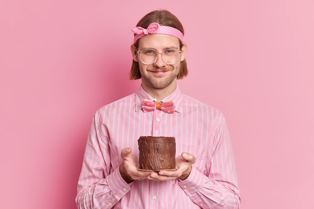 Un homme joyeux fête son anniversaire sur le lieu de travail reçoit les félicitations de ses collègues tient un petit gâteau avec une bougie allumée porte de grands lunettes et des vêtements de fête