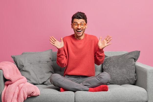 Un homme joyeux lève les paumes et réagit à une nouvelle impressionnante est assis les jambes croisées sur un canapé confortable