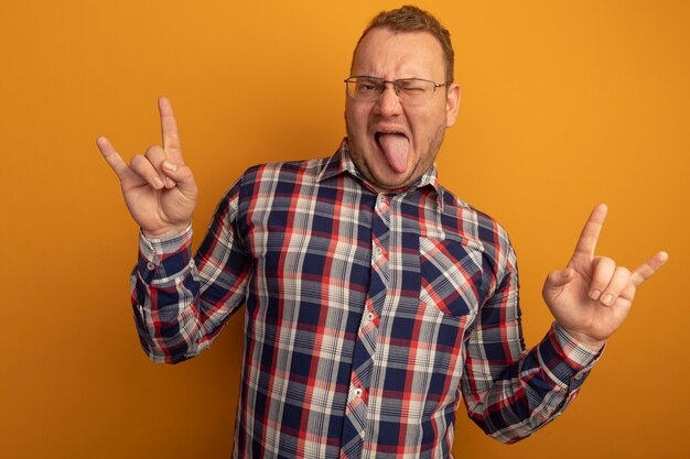 Homme joyeux à lunettes et chemise à carreaux heureux et joyeux montrant des symboles rock qui sort la langue debout sur un mur orange
