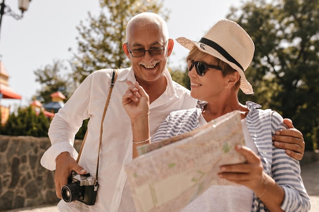 Homme joyeux avec moustache en tenue blanche avec caméra regardant la carte et souriant avec une femme aux cheveux courts en lunettes de soleil chapeau et chemise bleue en plein air