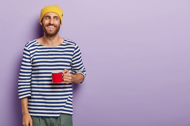 Un homme joyeux tient une tasse rouge, a une expression faciale heureuse, porte un chapeau jaune et un pull rayé