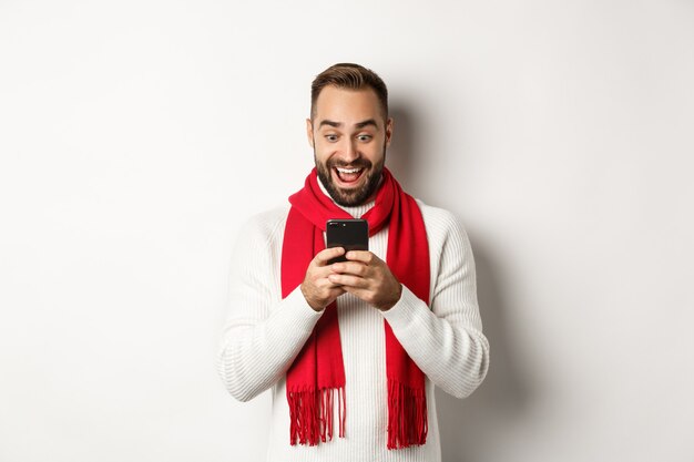 Homme lisant des messages sur son téléphone portable et ayant l'air heureux, debout dans un pull d'hiver et une écharpe rouge, fond blanc