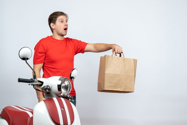 Homme de livraison incertain incertain en uniforme rouge debout près de scooter donnant une boîte de papier sur un mur blanc