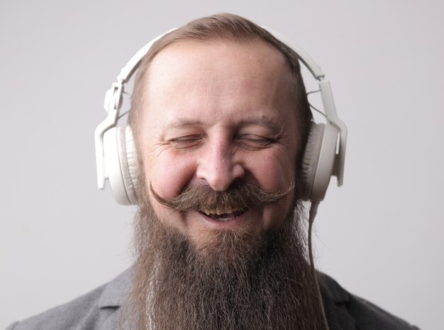 Homme avec une longue barbe et une moustache portant des écouteurs blancs, debout devant un mur gris