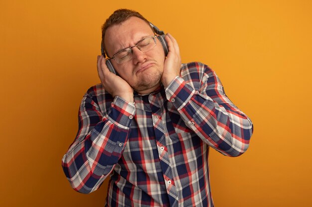 Homme à lunettes et chemise à carreaux avec un casque avec les yeux fermés appréciant sa musique préférée debout sur un mur orange