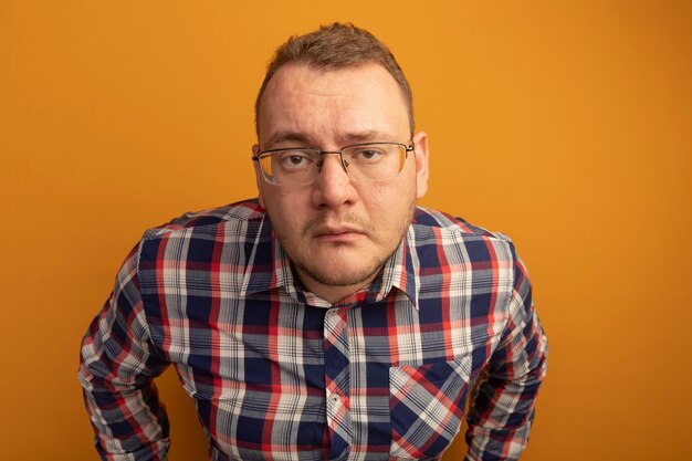 Homme à lunettes et chemise à carreaux avec expression triste debout sur un mur orange