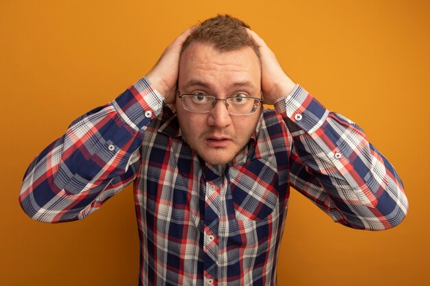 Homme à lunettes et chemise à carreaux inquiet et confus avec les mains sur la tête debout sur un mur orange