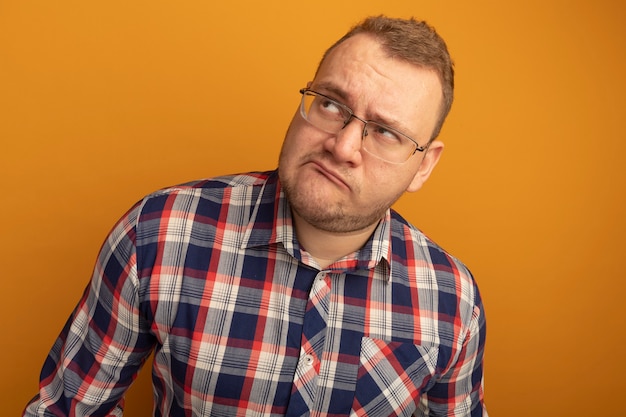 Homme à lunettes et chemise à carreaux lookign up perplexe debout sur un mur orange