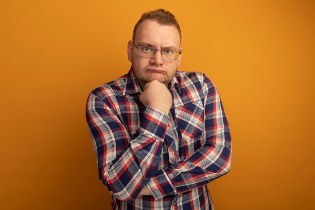 Homme à lunettes et chemise à carreaux avec la main sur son menton pensant avec un visage sérieux debout sur un mur orange