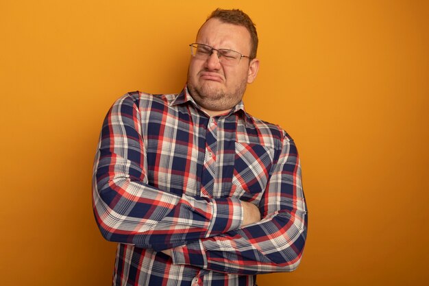 Homme à lunettes et chemise à carreaux mécontent de froncer les sourcils avec les bras croisés debout sur le mur orange