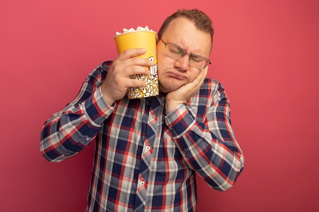 L'homme à lunettes et chemise à carreaux montrant seau avec du pop-corn avec la main sur son visage avec les yeux fermés s'ennuie debout sur le mur rose