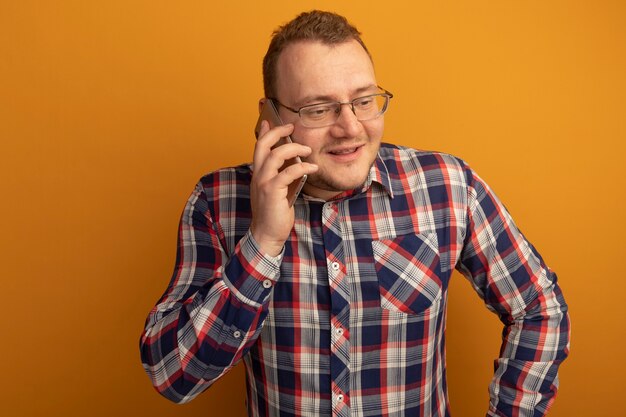Homme à lunettes et chemise à carreaux souriant tout en parlant au téléphone mobile debout sur un mur orange