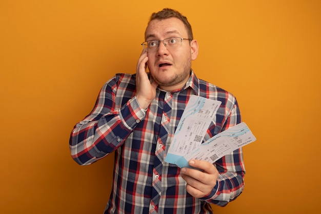 Homme à lunettes et chemise à carreaux tenant des billets d'avion à côté surpris et étonné debout sur un mur orange