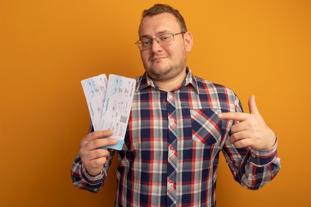 Homme à lunettes et chemise à carreaux tenant des billets d'avion pointant avec l'index sur eux souriant confiant debout sur le mur orange