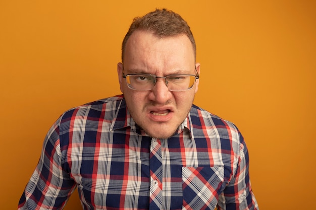 Homme à lunettes et chemise à carreaux avec visage en colère debout sur un mur orange