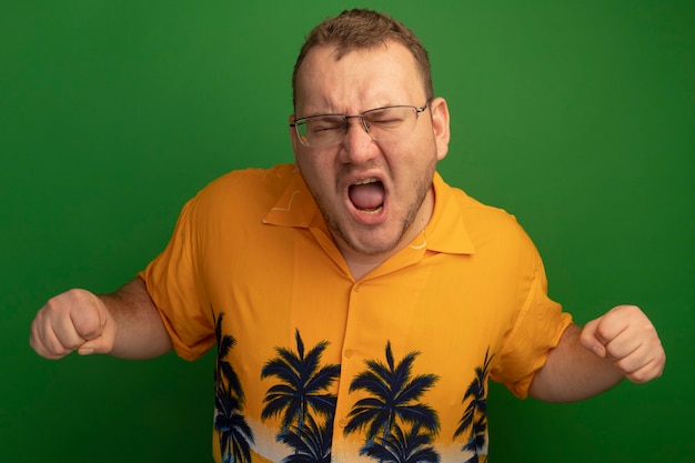 Photo gratuite homme à lunettes et chemise orange criant avec les poings serrés en colère et frustré debout sur le mur vert