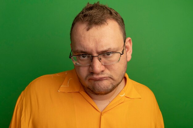 Homme à lunettes et chemise orange insatisfait des sourcils froncés debout sur le mur vert