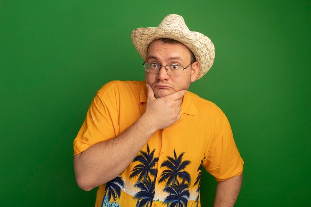 Homme à lunettes portant chemise orange et chapeau d'été perplexe debout sur le mur vert