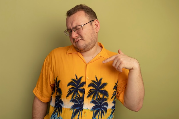 Homme à lunettes portant une chemise orange à la confusion et mécontent de pointer du doigt sur lui-même debout sur un mur léger