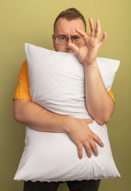 Homme à lunettes portant une chemise orange étreignant un oreiller montrant un geste de petite taille, symbole mesure debout sur mur léger