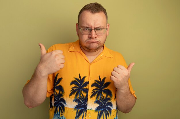 Homme à lunettes portant une chemise orange avec visage en colère fronçant les sourcils montrant les pouces vers le haut debout sur un mur léger