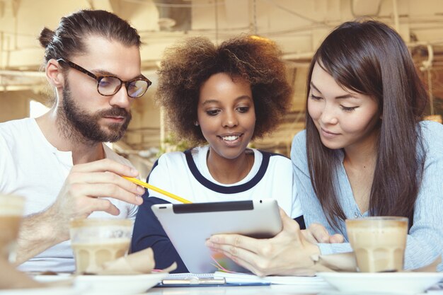 Homme à lunettes présentant la stratégie commerciale sur tablet pc, montrant des informations à l'écran avec un crayon pendant que ses collègues asiatiques et africains l'écoutent avec attention, assis à l'espace de travail collaboratif