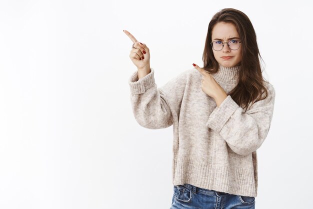 homme à lunettes et pull, fronçant les sourcils et pinçant les lèvres dans l'aversion et le doute en pointant vers le coin supérieur gauche, insatisfait du produit.