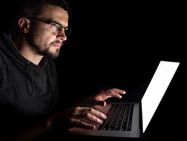 Un homme à lunettes regarde sérieusement un écran d'ordinateur portable, travaille sur un ordinateur la nuit dans le noir.