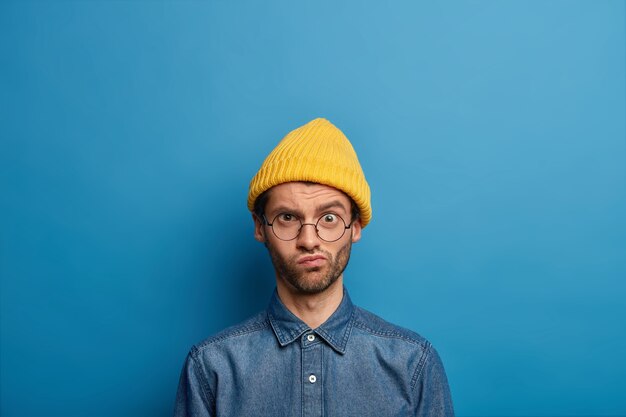 Homme mal rasé embarrassé regarde avec une expression de visage mécontent de froncement de sourcils à la caméra, porte des lunettes optiques, un chapeau jaune et une chemise en jean