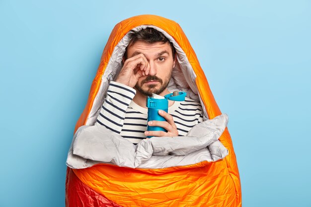 Un homme mal rasé de fatigue se frotte les yeux, a une expression endormie, voyage en camping, se détend dans un sac de couchage, profite d'un week-end d'aventure