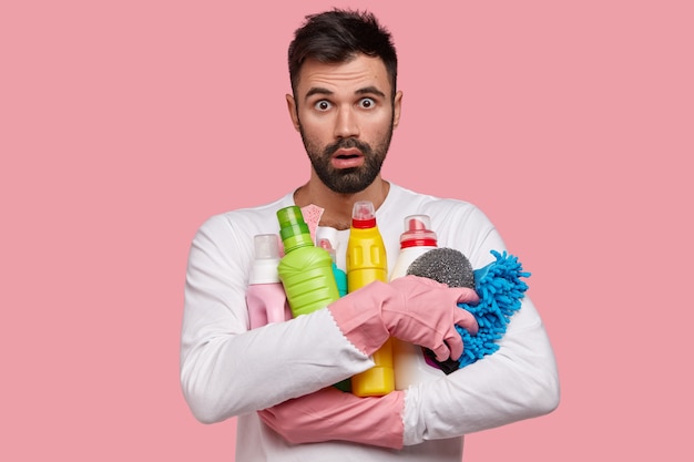 Photo gratuite un homme mal rasé de race blanche choqué porte des produits de nettoyage, une éponge pour laver la vaisselle, vêtu d'une tenue décontractée, regarde avec stupéfaction, fait les corvées