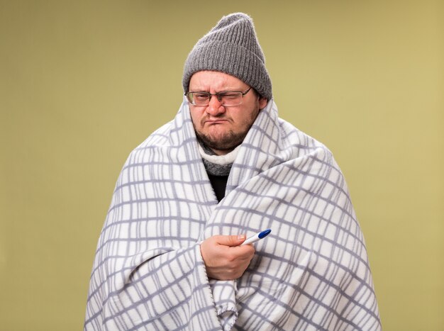 Homme malade d'âge moyen mécontent portant un chapeau d'hiver et une écharpe enveloppés dans un plaid tenant un thermomètre