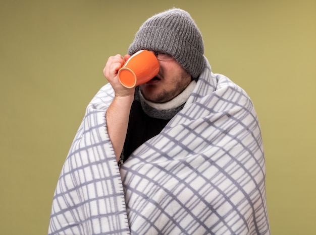 Un homme malade d'âge moyen portant un chapeau d'hiver et une écharpe enveloppés dans un plaid boit du thé dans une tasse isolée sur un mur vert olive