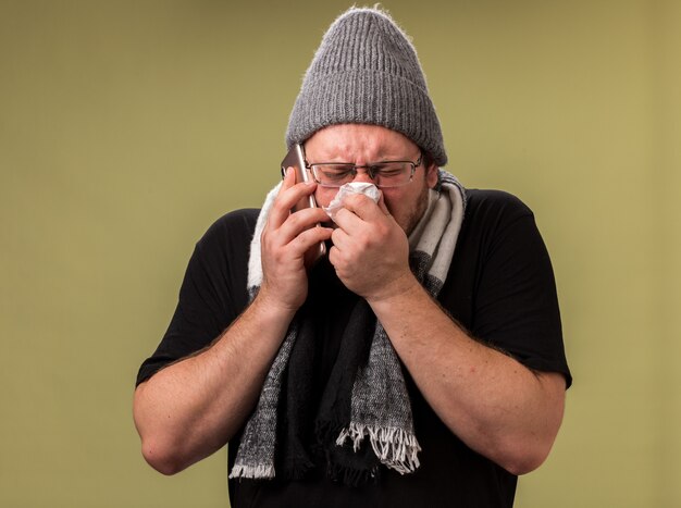 Un homme malade d'âge moyen portant un chapeau d'hiver et une écharpe parle au téléphone en essuyant le nez avec une serviette isolée sur un mur vert olive