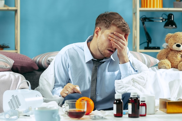 Photo gratuite homme malade barbu avec cheminée assis sur un canapé à la maison recouvert d'une couverture chaude et buvant du sirop contre la toux.