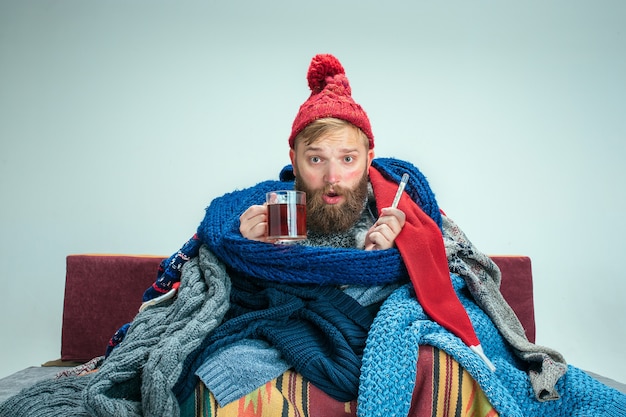 Homme malade barbu avec cheminée assis sur un canapé à la maison ou en studio avec une tasse de thé recouverte de vêtements chauds tricotés. Maladie, concept de grippe. Détente à la maison. Concepts de santé.