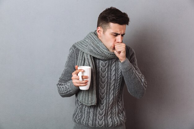 Homme malade en pull et écharpe tenant une tasse de thé tout en ayant la toux