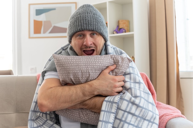 homme malade surpris avec une écharpe autour du cou portant un chapeau d'hiver enveloppé dans un plaid tenant un oreiller assis sur un canapé dans le salon