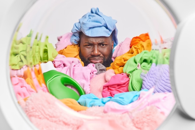 Un homme malheureux recouvert de linge multicolore charge une machine à laver avec des vêtements utilise un détergent liquide le visage ricane a un cavalier bleu sur la tête