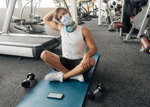 Homme avec masque médical et écouteurs à la salle de sport sur tapis