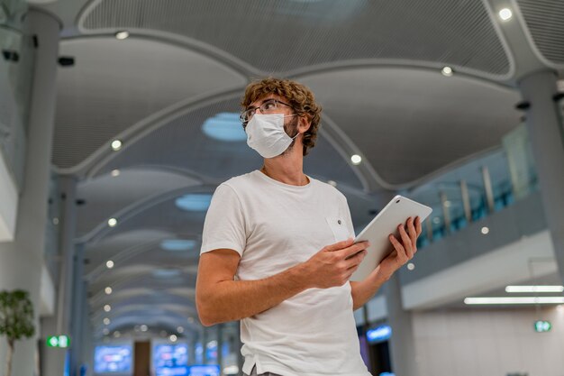 L'homme en masque respiratoire attend le prochain avion à l'aéroport et en utilisant une tablette.