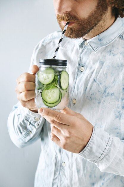 Homme méconnaissable en chemise de jeans léger boit du concombre frais fait maison avec de la limonade pétillante à la menthe à travers la paille rayée du pot transparent rustique dans les mains