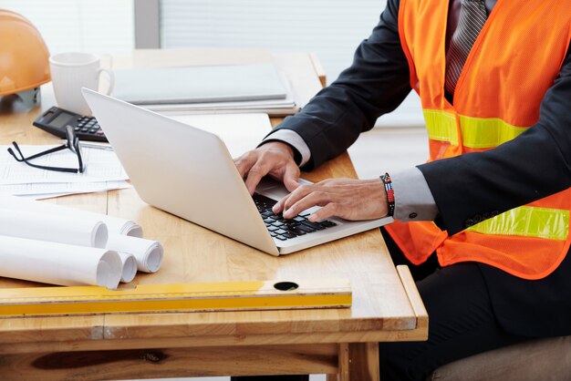 Homme méconnaissable en gilet de sécurité néon et tailleur assis au bureau et utilisant un ordinateur portable