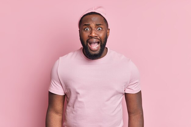 Un homme mécontent avec une barbe épaisse a une expression nerveuse inquiète s'exclame fortement garde la bouche ouverte porte un chapeau et un t-shirt décontracté pose contre un mur rose pâle